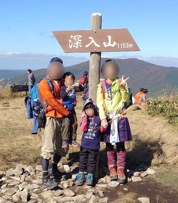 深入山の山頂標識（標高1153m）前で記念撮影する家族4人