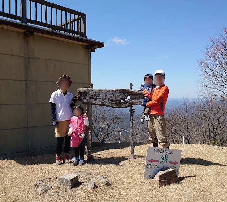 鷹ノ巣山の山頂標識の前に立つ家族4人。背後に休憩小屋と山並みが見える