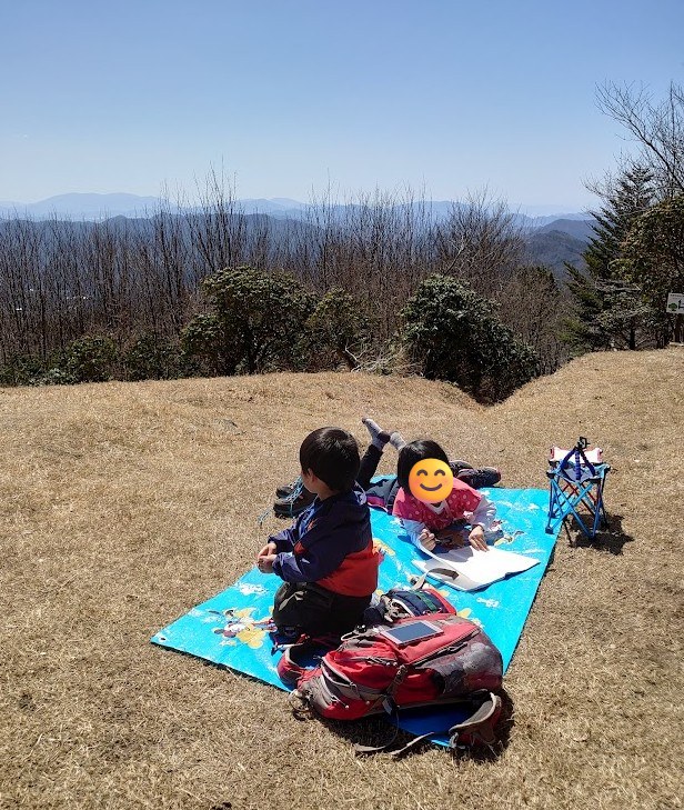 鷹ノ巣山の広い山頂で、レジャーシートに寝そべって記帳ノートに書き込む6歳の娘と、そばに座る3歳の息子