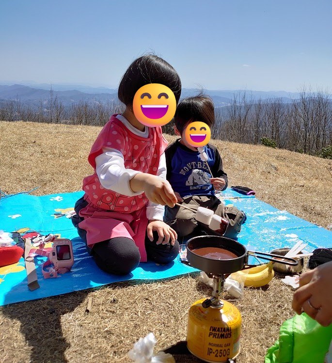 山頂でシングルバーナーを使いチョコレートフォンデュを楽しむ娘と息子。背景に山並みの展望