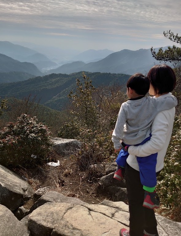 可部冠山の山頂の岩の上で、ママに抱き上げられて幾重にも重なる山並みを眺める3歳の息子。ふたりとも後ろ姿