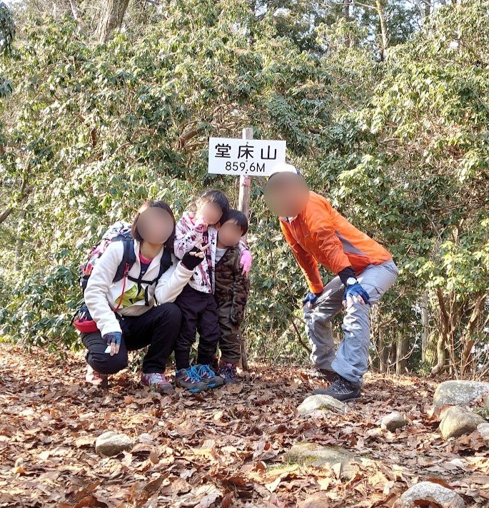 堂床山の山頂に到着し、山頂標識の前で喜ぶ家族4人