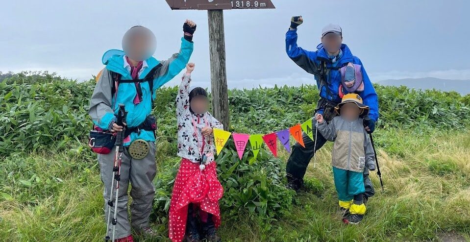 山頂標識の前で、こぶしを上げて全20座の完走を喜ぶ家族4人。「祝 里山キング」の手作りガーランドが飾られている