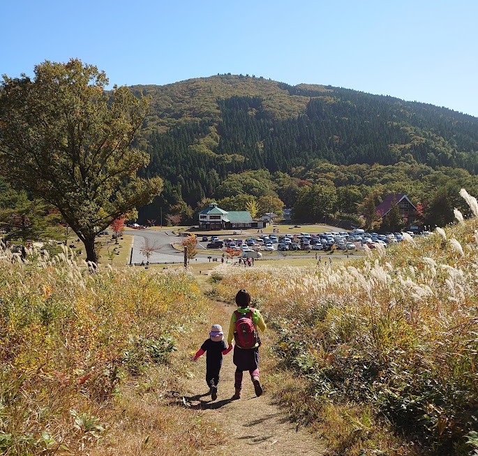 深入山のススキの間の道を歩く母と娘と、その先に見える登山口の駐車場